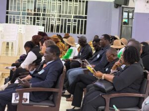 Audience seated in a conference hall, many reading pamphlets, some in black attire and colorful traditional outfits with hats, badges visible.