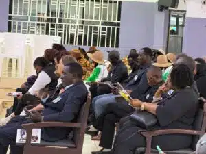 Audience seated in a conference hall, many reading pamphlets, some in black attire and colorful traditional outfits with hats, badges visible.