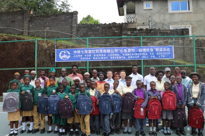 Group of students and adults posing outdoors with backpacks, in front of a banner about a Sierra Leone donation project in progress.