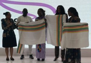 Five people stand on a stage presenting large white textiles with rainbow stripes, displaying the designs to the audience.