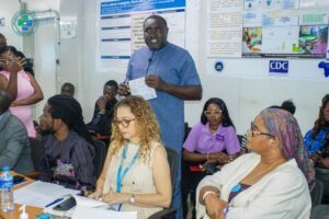 | The Calabash Newspaper Man in blue tunic standing and speaking at a conference, holding a note card, with posters on the wall behind him.