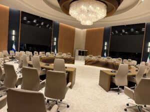 Empty modern boardroom with beige swivel chairs around U-shaped desks; flags in the corners and a grand chandelier overhead.