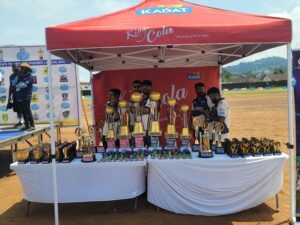 Trophies and medals arranged on a white-clothed table under a red tent at an outdoor sports event. Kadat branding visible on tent backdrop and banners in the background.