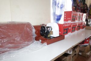 Row of red Singer sewing machine boxes on a display shelf with wrapped machines and fabric rolls in a shop setting.