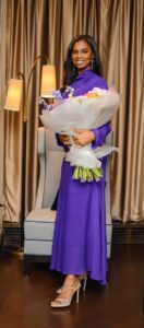 | The Calabash Newspaper Woman in a purple dress holding a large bouquet wrapped in white tissue, standing beside a blue chair in a softly lit room with curtains and a floor lamp.