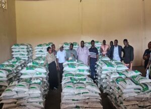 Group of people posing in a warehouse surrounded by tall stacks of white bags with green markings on pallets under a yellow wall.