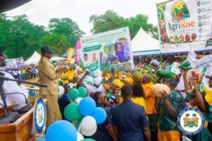 Crowd at an outdoor agricultural festival; a speaker addresses attendees from a podium amid colorful banners, tents, and balloons.