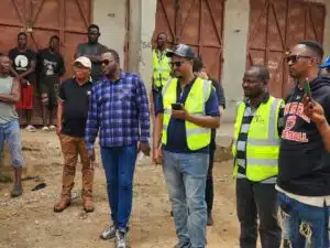 Group of men standing outdoors, several in neon safety vests, watching something off-camera.