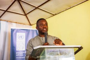 James B. Sandi delivering the keynote address at the Orientation Launch Ceremony | The Calabash Newspaper Man speaking into a handheld microphone at a lectern, with a UNDP banner in the background.