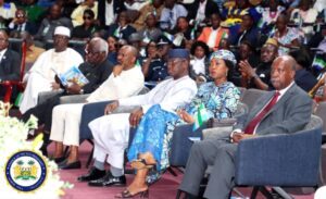 | The Calabash Newspaper Front row of dignitaries at a formal ceremony, including a man in white traditional attire, a woman in a blue patterned dress, and a suited man, with a Nigerian flag and official seal visible in the foreground.