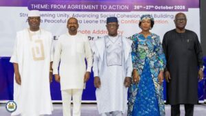 Five adults in traditional African clothing pose on a stage in front of a banner reading 'Theme: From Agreement to Action'.