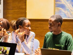 Three young adults sit at a table in a discussion, a woman gesturing as she talks to a man in a green shirt; laptops open.