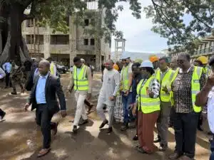 Group of people in high-visibility vests walking through a muddy construction site with an unfinished building nearby, trees overhead.