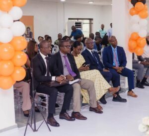 | The Calabash Newspaper Group of professionally dressed people seated in rows at a formal event, orange and white balloon decorations along columns in a bright hall.