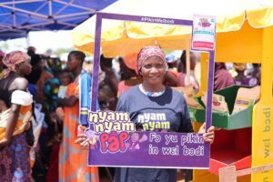 Woman in a headscarf holds a purple Nyam Nyam poster at a bright market stand, with people in the background.