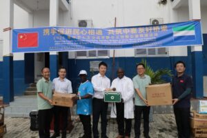 Group of people standing under a blue banner, handing over boxes in a donation event between China and Sierra Leone teams.