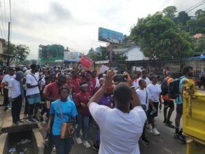 Crowd of people marching down a city street; many wear colored shirts while a man in white films them with a smartphone.