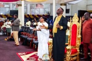 Religious ceremony with people standing and praying, a woman in white and a man in black/gold robes near a gold throne in a decorated hall.