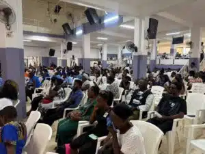 Large hall filled with diverse attendees seated in white plastic chairs, listening to a presentation at a conference or meeting session.