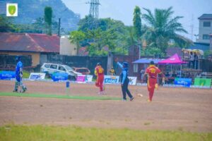 | The Calabash Newspaper Cricket field during a match with players in red and blue uniforms and an umpire signaling near the wicket fence lined with sponsor banners.