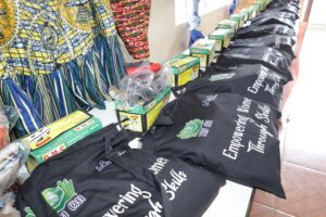 | The Calabash Newspaper Market stall displaying GMC charcoal iron boxes lined up on a table, with black bags reading 'Empowering Women' and bright patterned fabrics in the background.