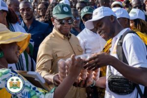 | The Calabash Newspaper Man in tan uniform and green cap hands out loaves of bread to a smiling young man wearing a white cap at a community bread distribution event, with a crowd and a seal/logo visible in the corner.