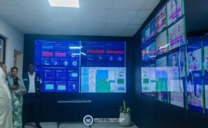 Group of people standing in a high-tech operations room with wall-sized screens displaying dashboards, charts, and a map; ministry of technology and innovation logo at the front.