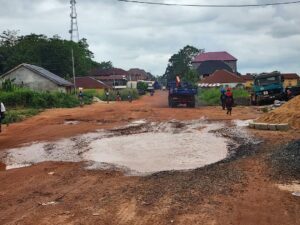 Unpaved red dirt road with a large muddy puddle, residential buildings, and heavy trucks in a rural construction area in the distance.