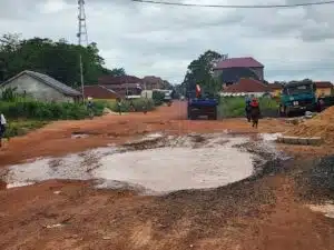 Unpaved red dirt road with a large muddy puddle, residential buildings, and heavy trucks in a rural construction area in the distance.