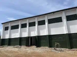 White building with a dark green base, tall vertical columns, and rectangular lattice vents along the windows; bare dirt foreground suggests construction.