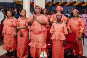 | The Calabash Newspaper Group of women in peach lace dresses and head wraps standing and clapping at a church service, with decorated pillars and red chairs in the background.