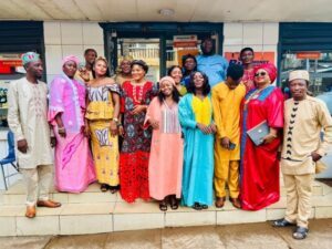 Group of men and women in colorful traditional African clothing posing on steps outside a storefront, smiling at the camera.