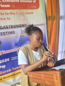 | The Calabash Newspaper Woman with braided hair speaking into a microphone at a wooden podium, in front of a colorful event banner.
