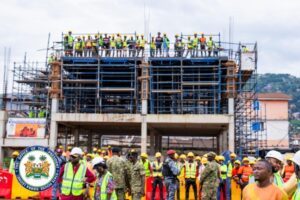 | The Calabash Newspaper Construction site with a multi-story frame under scaffolding; workers in hard hats and vests on top and along the ground, activelyBuilding.