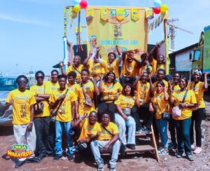 | The Calabash Newspaper Group of smiling people in yellow shirts posing for a festival photo with a colorful stage banner reading World Food Day and 'Onga Waka Fiesta' in the background